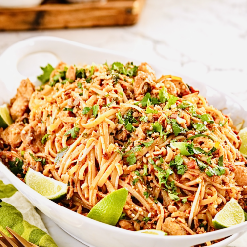 chicken pad thai noodles with lime wedges, crushed peanuts, cilantro, and golden fork on white serving platter
