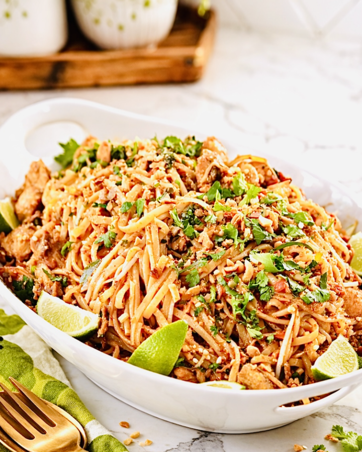 chicken pad thai noodles with lime wedges, crushed peanuts, cilantro, and golden fork on white serving platter
