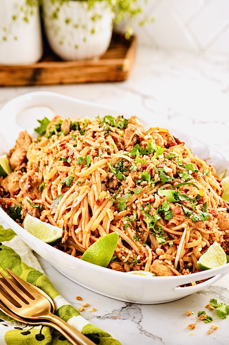chicken pad thai noodles with lime wedges, crushed peanuts, cilantro, and golden fork on white serving platter