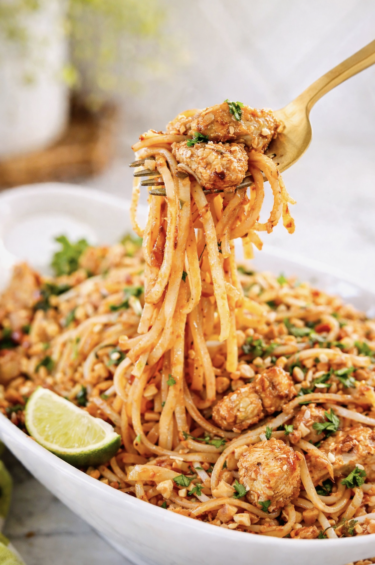 gold fork lifting glossy chicken pad thai noodles with peanuts and cilantro from serving dish