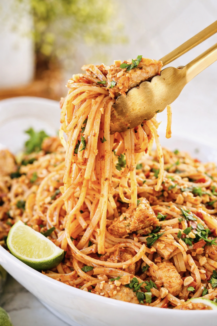 gold tongs lifting saucy chicken pad thai noodles with peanuts and cilantro from platter