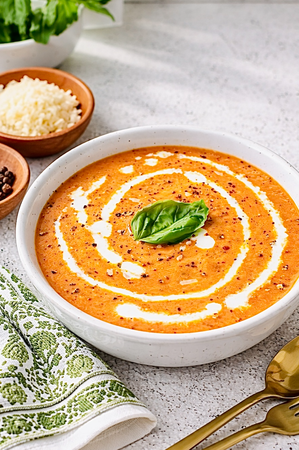 Creamy tomato soup garnished with a swirl of cream, cracked black pepper, and a fresh basil leaf, served in a white bowl on a light kitchen countertop with gold utensils.