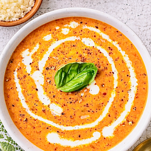 Creamy tomato soup garnished with a swirl of cream, cracked black pepper, and a fresh basil leaf, served in a white bowl on a light kitchen countertop with gold utensils.