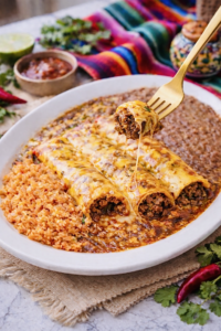 Tex-Mex beef enchiladas with chili gravy served with Mexican rice and refried beans, topped with melted cheddar cheese and a fork lifting a bite