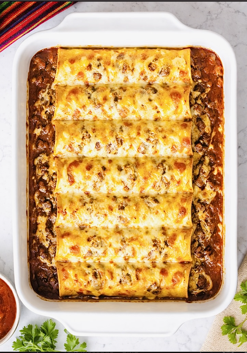 Baked Tex-Mex beef enchiladas in a white casserole dish covered with chili gravy and melted cheddar cheese
