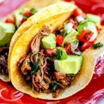 A close-up of tender shredded beef tacos served in warm corn tortillas, topped with fresh avocado cubes and vibrant pico de gallo, garnished with cilantro and lime wedges.