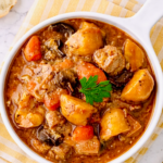 Overhead view of beef stew in a white bowl, showing chunks of beef, potatoes, carrots, mushrooms, and rich gravy, garnished with a parsley leaf and placed on a yellow cloth.