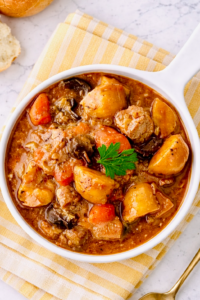 Overhead view of beef stew in a white bowl, showing chunks of beef, potatoes, carrots, mushrooms, and rich gravy, garnished with a parsley leaf and placed on a yellow cloth.