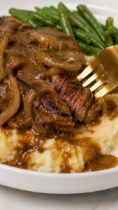 Fork cutting into fork-tender smothered steak with onion gravy over creamy mashed potatoes, served with green beans.