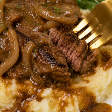 Fork cutting into fork-tender smothered steak with onion gravy over creamy mashed potatoes, served with green beans.