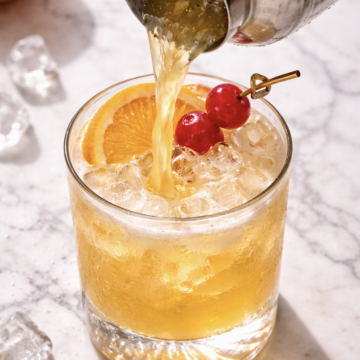 Whiskey sour being poured from cocktail shaker into rocks glass with fresh ice, orange slice, and maraschino cherries