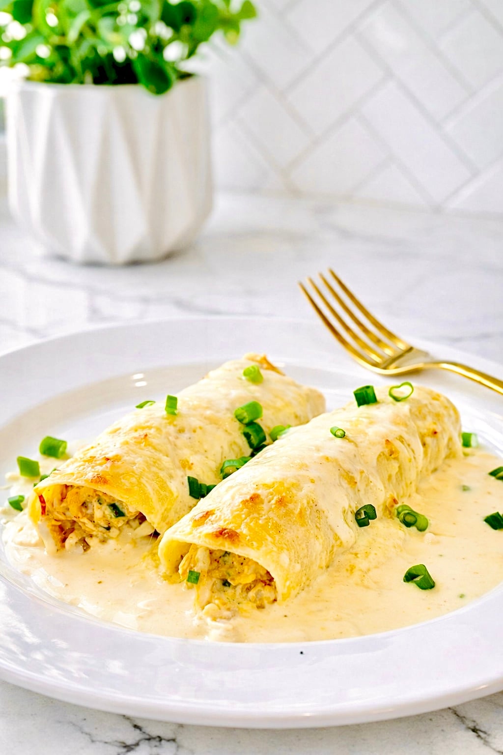 Creamy white chicken enchiladas topped with salsa verde sauce on a white plate with gold fork and bright kitchen background