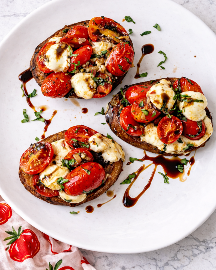 Tomato bruschetta with fresh mozzarella and balsamic glaze on toasted baguette slices, topped with cherry tomatoes and basil on a white plate