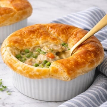 Chicken pot pie with flaky puff pastry crust in a white ramekin, creamy chicken and vegetable filling visible with a spoon inserted, set on a white marble-style countertop with a striped linen nearby.