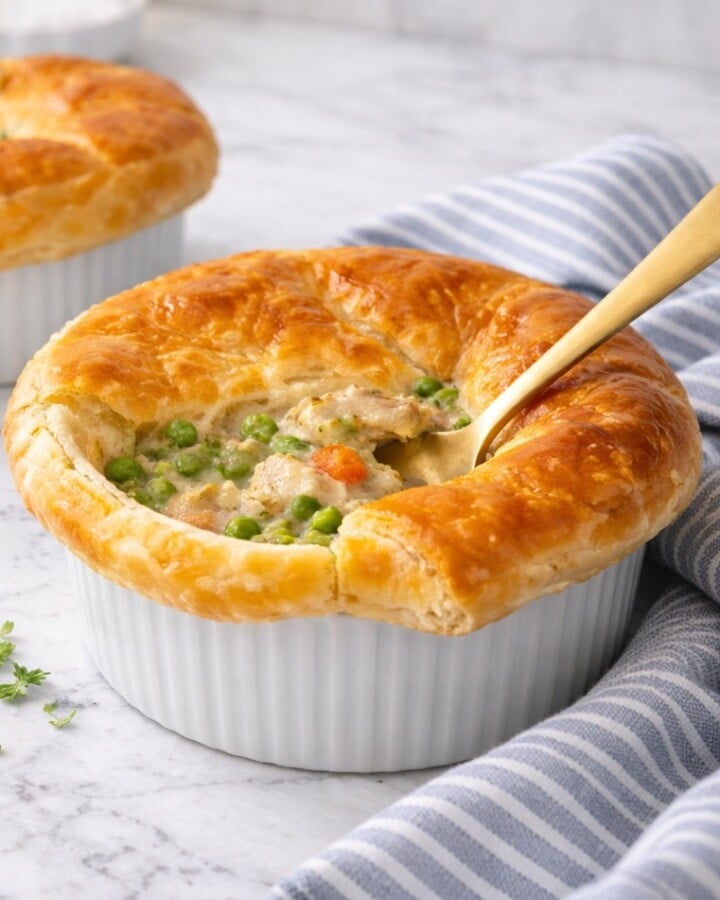 Chicken pot pie with flaky puff pastry crust in a white ramekin, creamy chicken and vegetable filling visible with a spoon inserted, set on a white marble-style countertop with a striped linen nearby.