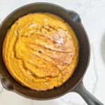 Freshly baked pumpkin spice cornbread in a cast iron skillet, featuring pumpkin purée, maple syrup, and autumn spices with a golden brown crust, perfect for fall meals.