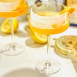 An elegant Sidecar cocktail served in a coupe glass with a sugared rim, featuring a vibrant orange twist garnish resting on the rim. The drink glows with a golden hue, hinting at the mix of cognac, Cointreau, and lemon juice inside.
