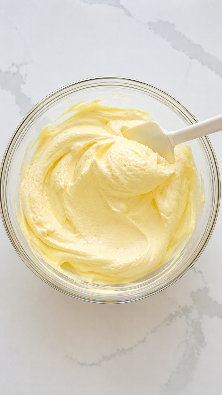 Creamy vanilla pudding mixture in a glass mixing bowl with a white spatula on a white quartz countertop for Biscoff banana pudding preparation.