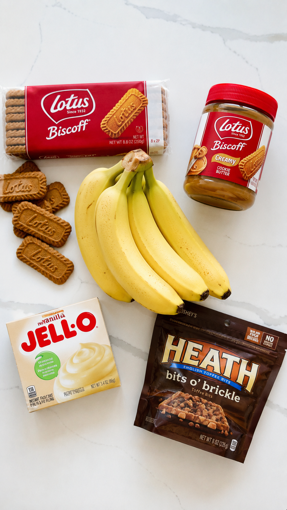 Ingredients for Biscoff banana pudding including bananas, vanilla pudding mix, Biscoff cookies, cookie butter, and toffee bits on a white quartz countertop.