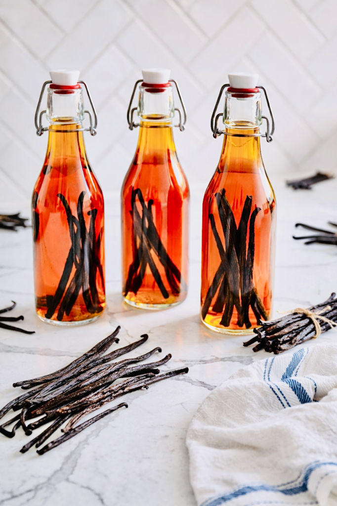 Three glass swing-top bottles filled with amber homemade vanilla extract and whole vanilla beans on a marble countertop with loose vanilla pods and a striped kitchen towel.