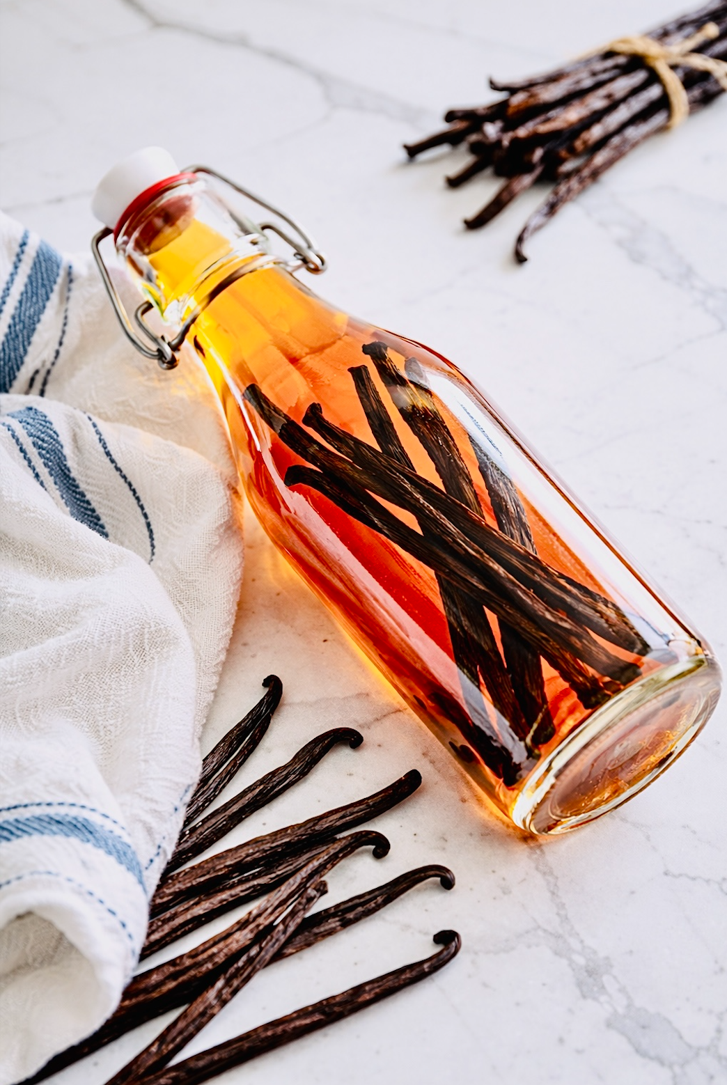 Glass bottle of homemade vanilla extract with whole vanilla beans inside, resting on a marble surface beside scattered vanilla pods and a white towel with blue stripes.