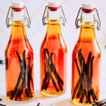 Three glass swing-top bottles filled with amber homemade vanilla extract and whole vanilla beans on a marble countertop with loose vanilla pods and a striped kitchen towel.