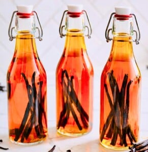Three glass swing-top bottles filled with amber homemade vanilla extract and whole vanilla beans on a marble countertop with loose vanilla pods and a striped kitchen towel.