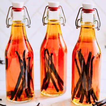 Three glass swing-top bottles filled with amber homemade vanilla extract and whole vanilla beans on a marble countertop with loose vanilla pods and a striped kitchen towel.
