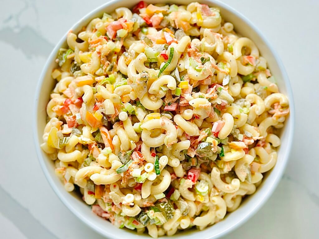 Bowl of creamy macaroni salad with elbow pasta, red peppers, celery, and chopped eggs, garnished with green onions and served on a rustic picnic table—perfect for summer potlucks and backyard BBQs.