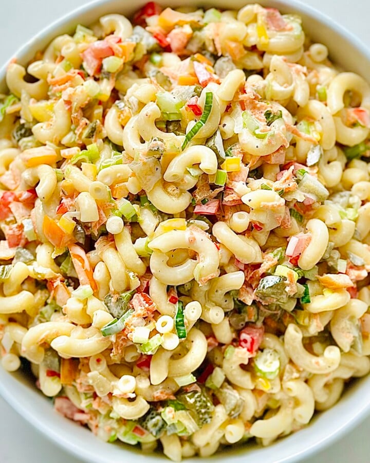Bowl of creamy macaroni salad with elbow pasta, red peppers, celery, and chopped eggs, garnished with green onions and served on a rustic picnic table—perfect for summer potlucks and backyard BBQs.