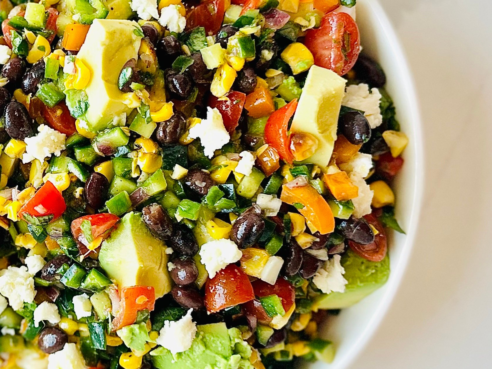 Fresh avocado, corn, black beans, and tomatoes mixed into a Mexican chopped salad in a white serving bowl
