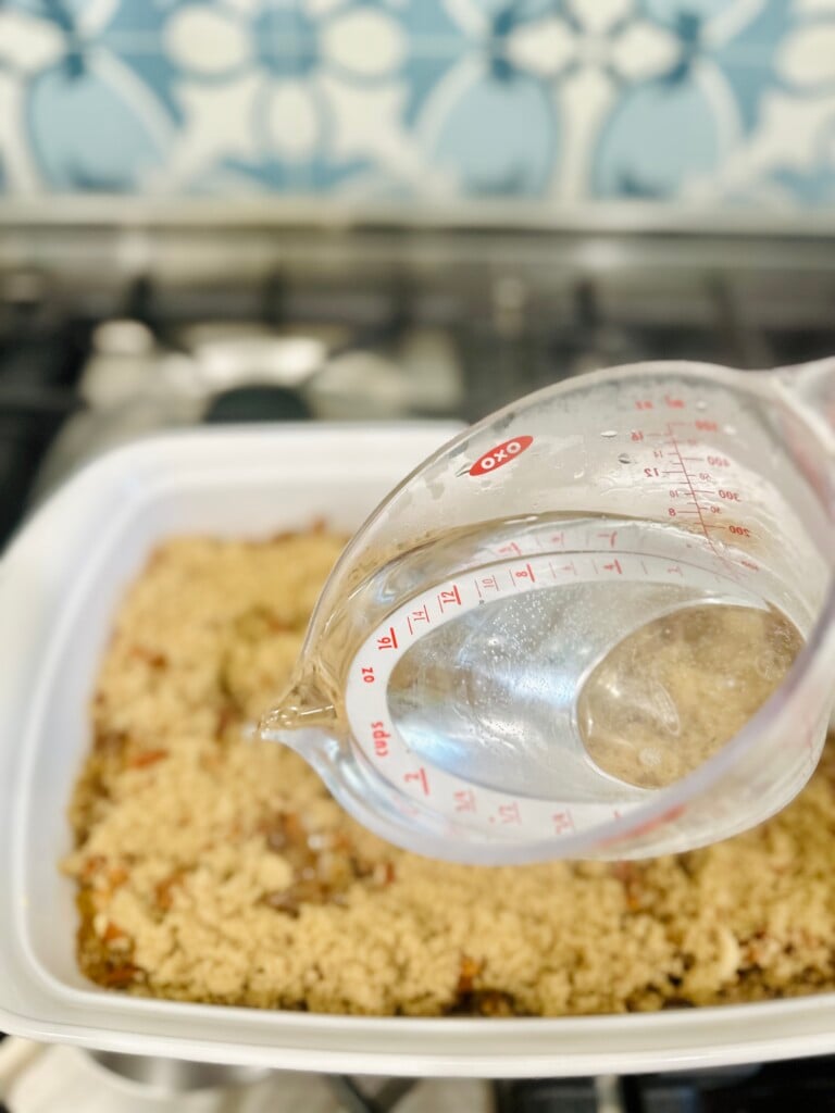 Boiling water being poured from a measuring cup over brown sugar and pecan layers in cobbler dish.