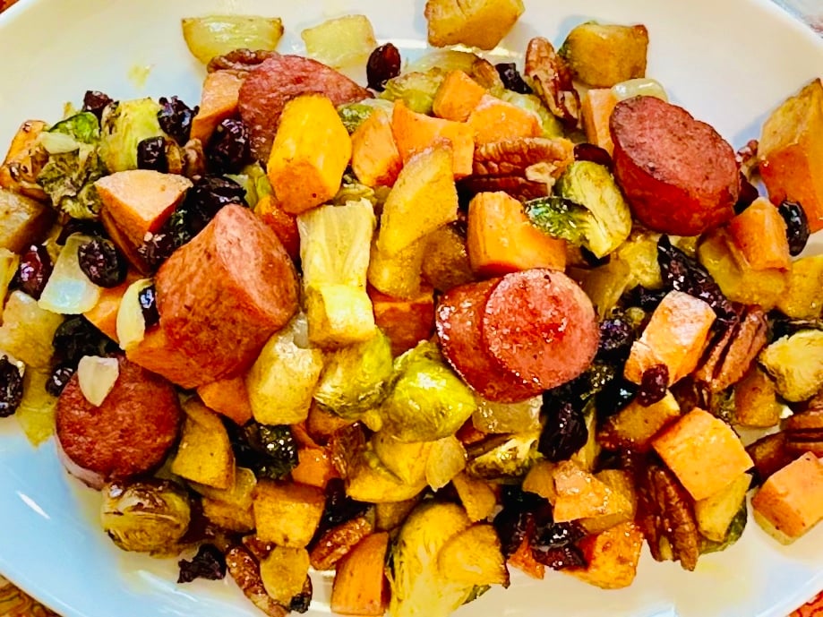 Overhead view of roasted sausage, sweet potatoes, apples, and Brussels sprouts with cranberries and pecans on a white serving platter.