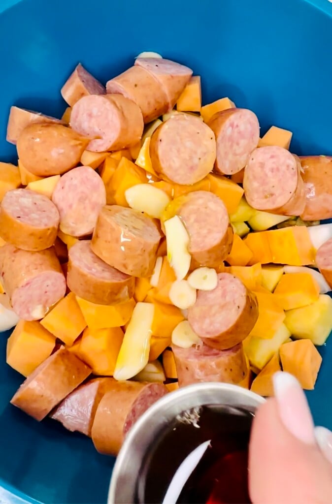 Maple syrup being added to chopped sausage, sweet potatoes, garlic, and Brussels sprouts in a blue mixing bowl before roasting.