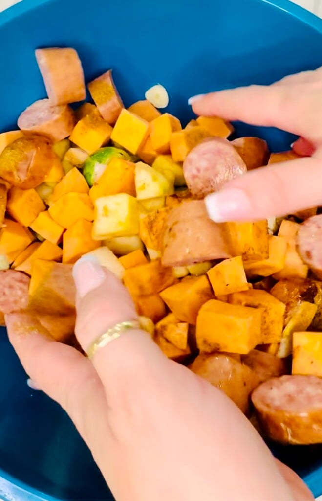 Hands tossing sausage, Brussels sprouts, and sweet potatoes together with seasoning and oil in a large mixing bowl.
