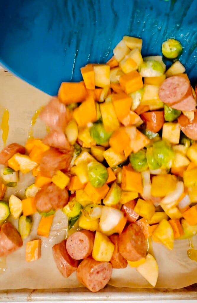 Seasoned sausage, Brussels sprouts, apples, and sweet potatoes being poured from a blue bowl onto a parchment-lined sheet pan.
