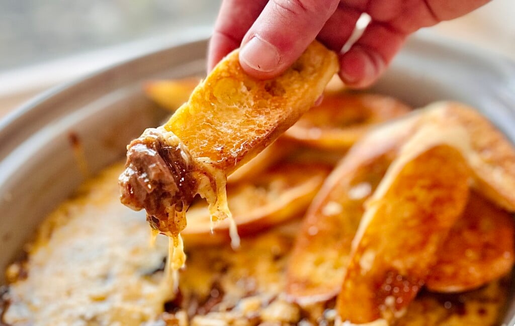 Baguette slice lifted from the pot showing tender beef strands and melted Gruyère stretching.
