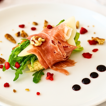 Close-up of prosciutto wrapped pear slices on arugula with balsamic reduction and festive toppings.