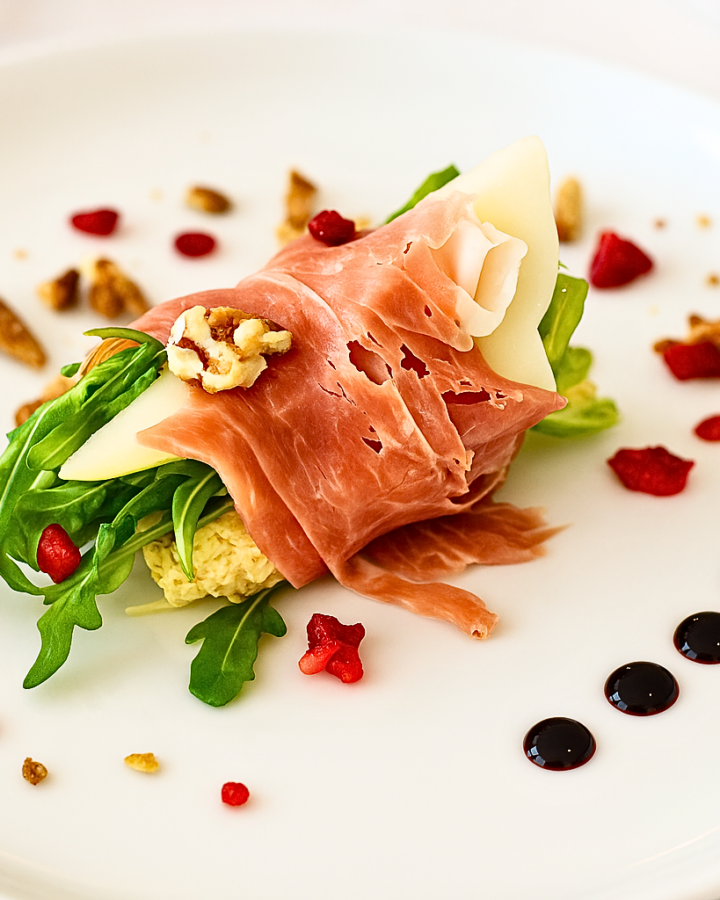 Close-up of prosciutto wrapped pear slices on arugula with balsamic reduction and festive toppings.