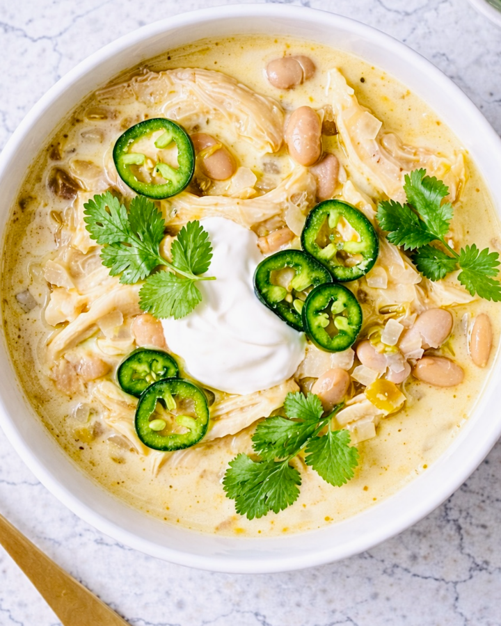 Creamy white chicken chili in a white ceramic bowl, topped with sliced jalapeños, fresh cilantro, and a dollop of sour cream, photographed from overhead on a light marble surface.