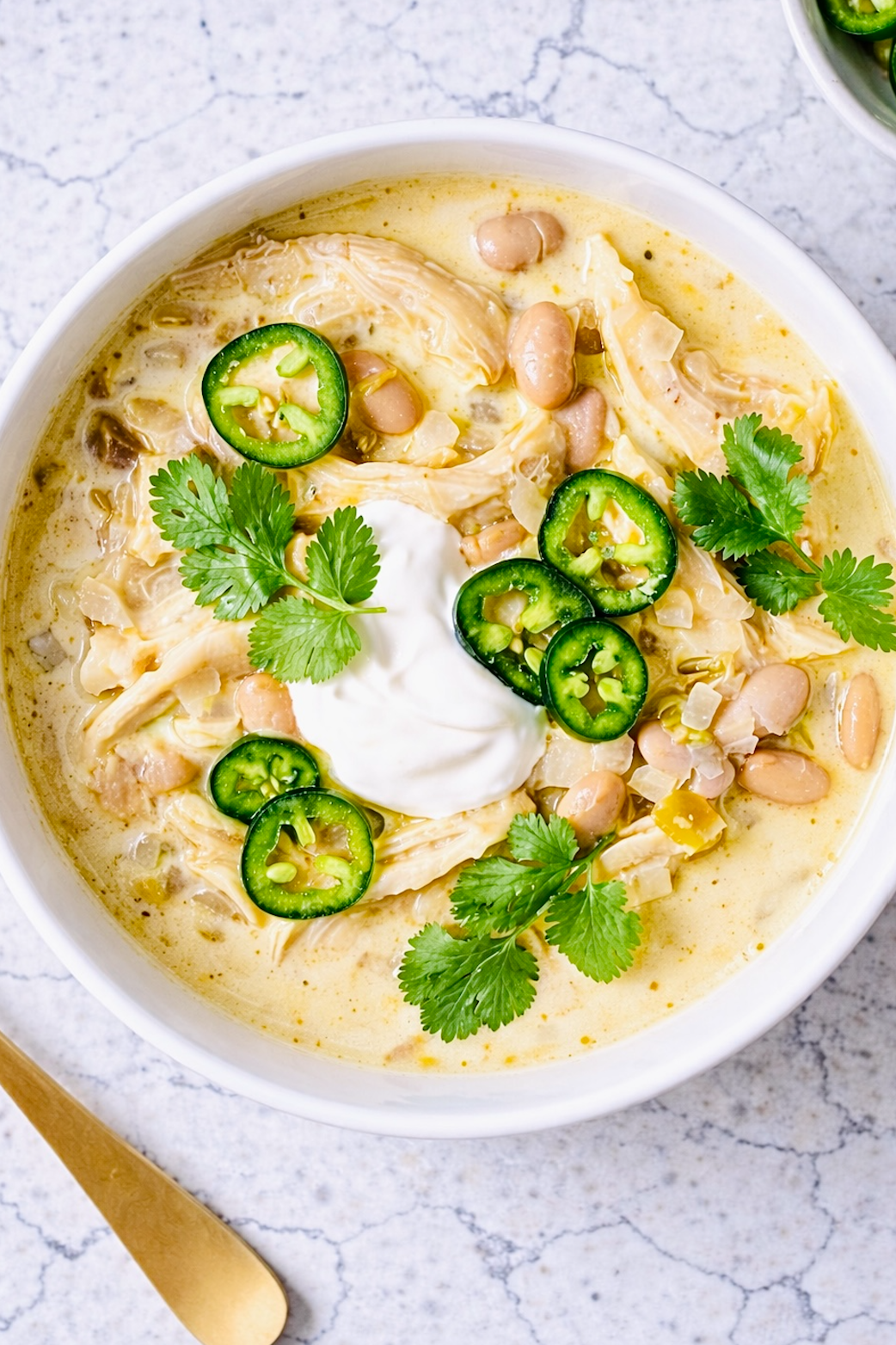 Creamy white chicken chili in a white ceramic bowl, topped with sliced jalapeños, fresh cilantro, and a dollop of sour cream, photographed from overhead on a light marble surface.