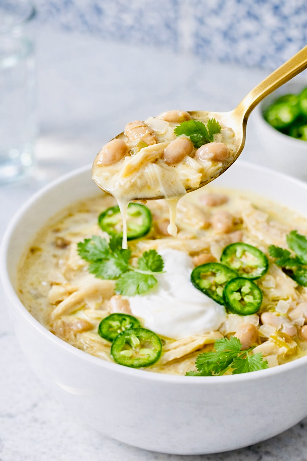 Spoon lifting creamy white chicken chili with shredded chicken and white beans, showing melted texture and steam, with jalapeños and cilantro visible in the bowl below.