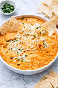 Buffalo chicken dip in a white round baking dish with melted cheese, blue cheese crumbles, and green onions, served with tortilla chips on a marble surface.