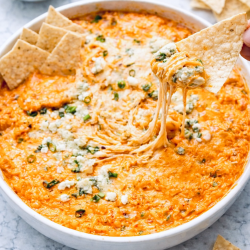 Buffalo chicken dip in a white round baking dish with melted cheese, blue cheese crumbles, and green onions, served with tortilla chips on a marble surface.