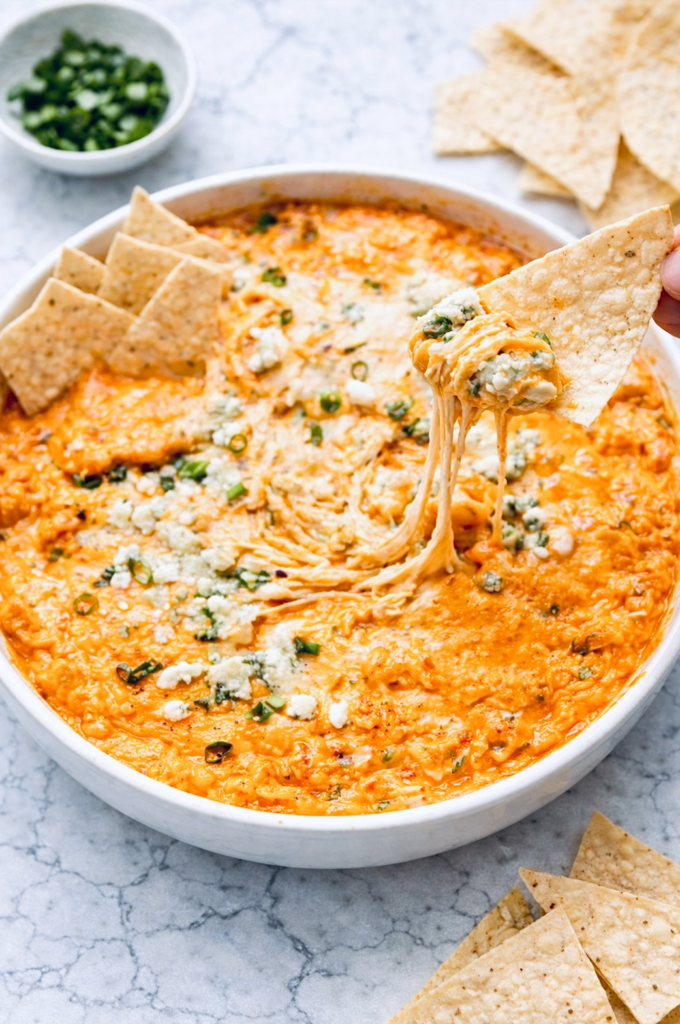 Buffalo chicken dip in a white round baking dish with melted cheese, blue cheese crumbles, and green onions, served with tortilla chips on a marble surface.