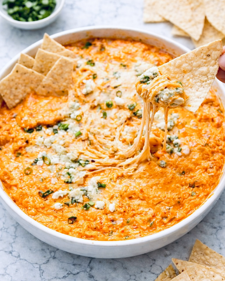 Buffalo chicken dip in a white round baking dish with melted cheese, blue cheese crumbles, and green onions, served with tortilla chips on a marble surface.