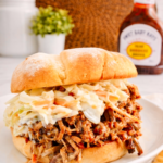 Pulled pork sandwich on a soft bun topped with creamy coleslaw, served on a white plate in a bright kitchen setting