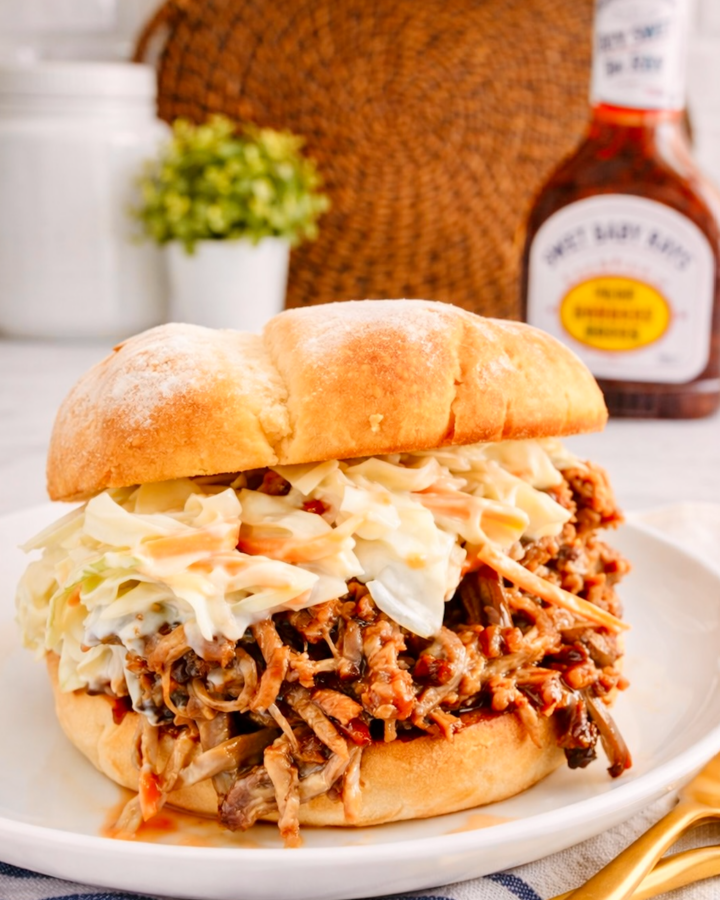Pulled pork sandwich on a soft bun topped with creamy coleslaw, served on a white plate in a bright kitchen setting