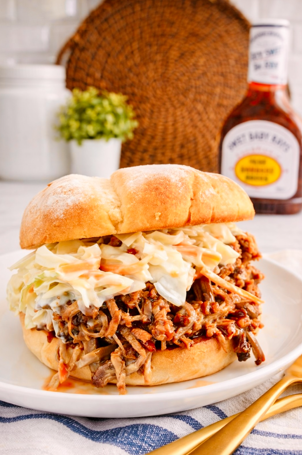 Pulled pork sandwich on a soft bun topped with creamy coleslaw, served on a white plate in a bright kitchen setting