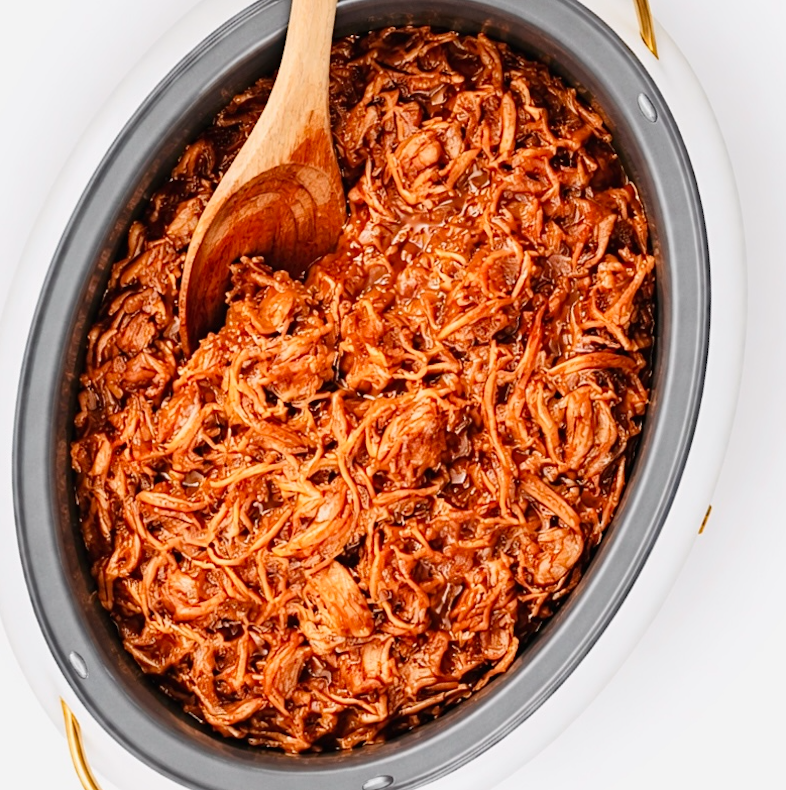 Shredded barbecue pork stirred inside a white slow cooker with a wooden spoon, showing tender texture and sauce